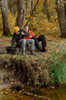 © m-art - Two friends relax by a serene riverbank, casting fishing lines into the calm water. Surrounded by vibrant autumn leaves, they share laughter and stories under the clear sky.