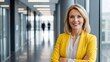 © Desingora - Portrait of a confident, smiling blonde businesswoman in a yellow blazer standing in a modern office hallway
