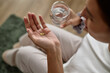 © Seventyfour - Young adult Caucasian woman sitting and holding two pills in one hand while holding glass of water in other hand, preparing to take medication, partial face visible