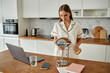 © Seventyfour - Caucasian young adult woman standing in modern kitchen holding round mirror and hairbrush preparing for morning routine with laptop, smartphone, notebook and glass of water on wooden counter