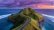 © liliyabatyrova - A winding road snakes up a grassy hill towards a summit overlooking the shimmering ocean. The sky above bursts with vivid purple and orange hues as the sun begins to set