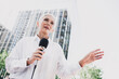 © deagreez - Confident elderly woman in formalwear speaks into a microphone outdoors in a modern city center about leadership and community