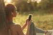 © SHOTPRIME STUDIO - A young woman enjoys music on her smartphone while sitting outdoors under a tree, dressed in casual sporty attire, conveying relaxation and tranquility
