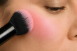 © propiks - A woman's face with a makeup brush applying pink blush to her cheek, showcasing a natural makeup look with a focus on enhancing facial features.
