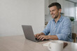 © Jeannette - Smiling man working remotely on laptop with earbuds in modern home office