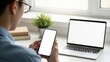 © onlyAW - Person holding smartphone next to laptop on desk by window with books and plant