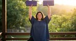 © Image - A woman smiles and holds yoga blocks overhead on a deck, backlit by the morning sun.