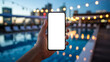 © Art_spiral - First person point of view of a hand holding a smartphone with a blank white mockup screen at a luxury rooftop swimming pool with blurred city skyline and warm evening lights