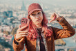 © EdNurg - Young woman with pink hair showing disapproval, thumbs down, while taking a selfie with smartphone in urban city