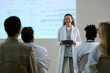 © AnnaStills - Caucasian young adult woman presenting medical data on digital tablet to diverse group of healthcare professionals during conference, standing in front of projected scientific charts