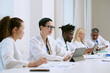 © AnnaStills - Group of multiethnic middle aged and young adult men and women wearing lab coats, working together at table using digital tablets and documents during scientific meeting