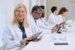 © AnnaStills - Caucasian senior woman smiling while holding digital tablet, sitting at desk with diverse group of middle aged and young adult scientists working with documents and laptops