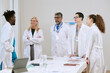 © AnnaStills - Group of middle aged and senior doctors including Black man, Caucasian woman, South Asian man, Caucasian woman and young adult Caucasian woman standing and discussing in medical office