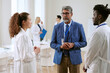 © AnnaStills - Middle aged Caucasian man with gray hair and beard wearing glasses and blue jacket, talking with young adult Caucasian woman and young adult Black man in medical uniforms in conference room