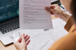 © pressmaster - Young adult woman holding contract document while reviewing paperwork at desk, laptop with data charts visible in background, hands and partial profile in focus