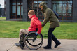 © Alex - A man in a wheelchair is riding on the sidewalk while a woman is helping him.