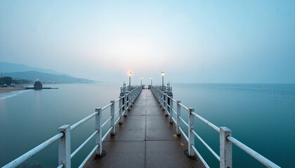  Symmetrical pier extending into misty blue ocean at dawn. Empty jetty with street lamps and vanishing point perspective
