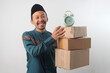 © Jamaludinyusup - Happy Asian Muslim man wearing a green traditional outfit and black songkok, smiling at the camera while holding a stack of cardboard boxes with Alarm Clock on Top. Isolated on a white background