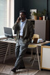 © DragonImages - Serious Black man standing in modern office talking on smartphone, leaning against desk with computer monitor and office supplies near window blinds