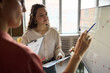 © DragonImages - Young adult Caucasian woman listening attentively while colleague explaining data on whiteboard during business meeting in modern office, both holding documents and discussing strategy