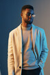 © SHOTPRIME STUDIO - Stylish African American man in a trendy beige coat and sunglasses, expressing confidence against a blue and gray gradient background