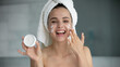 © fizkes - Effective skincare formula. Head shot portrait excited young woman with towel around head look at camera hold moisturizing cream bottle apply on face after shower advertize treatment cosmetic product