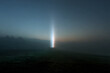 © kalafoto - A vertical beam of blue light piercing through dense fog in a dark mountain landscape under the night sky.