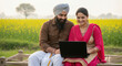 © Shivani - A rural Indian couple sits together on a charpai in a mustard field, using a laptop to access digital services amidst the countryside.