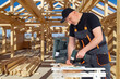 © Grispb - Construction, wooden house building. Carpenter assembles timber frame using a power drill, following plans, performing structural installation and residential construction work.