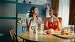 © stockbusters - Two besties holding cups with hot tea sitting together at apartment kitchen.