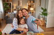 © Stockphotodirectors - A joyful family shares a light moment in a comfortable living room. Parents and their child smile happily as they capture a selfie with breakfast bowls nearby.