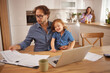 © Stockphotodirectors - A man sits at a table with paperwork and a laptop while holding a smiling child. A woman stands in the background, creating a warm, family-friendly atmosphere in a modern kitchen.