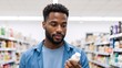 © Vasiliy - A young Black man shopping in a store and reading the label on a product. Thoughtful male consumer choosing personal care items in a supermarket aisle