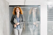 © Novak - Smiling business woman with orange curly hair leaving office building with laptop.