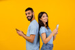 © Prostock-studio - A modern Arab couple stands back to back, smiling as they use their mobile phones. They are engaged with their devices, enjoying online interactions, in a vibrant yellow studio setting.
