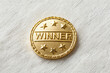 © lemonmoon - Shiny gold winner pin badge represents joyful victory success achievement. Round metal object symbolizes pride competition reward. Triumphant first place champion celebration on white texture