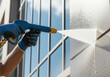 © Bakha - Worker using high pressure water jet to clean modern building facade, closeup of professional exterior cleaning on sunny day with protective gloves and reflective windows