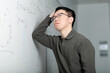© fotolite - A Chinese college student scratches his head in confusion while looking at math equations on a whiteboard. He is stuck and frustrated while trying to solve a challenging math problem in the classroom.