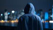 © Curioso.Photography - Person in a hoodie standing against a city skyline at night creating a moody urban solitude scene.