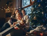 Mała, szczęśliwa dziewczynka wyjmująca lalkę z pudełka którą otrzymała jako prezent pod choinkę. W tle widać rodzinę która obserwuje jej radość
