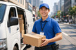 © imagemir - Smiling Asian male delivery driver holding a package beside his delivery van in an urban setting.