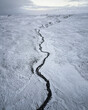 © AmazingAerialAgency - Aerial view of a dark river snaking through a vast, snow-covered landscape under a muted sky, creating a stark contrast between the dark water and the white expanse., Vik, Western Region, Iceland.