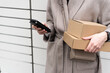 © Natali - A person is standing in front of mailboxes while holding two brown cardboard boxes and a phone. The scene shows a typical delivery activity in a city environment.