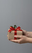 © josemperal - Manos sosteniendo regalo de Navidad rústico con lazo rojo sobre fondo gris con espacio de copia