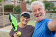 © Daniel - Old senior couple outdoors in paddle tennis court smiling and looking at the camera taking a selfie picture together playing padel. Active mature people concept