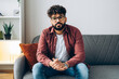 © Maria Vitkovska - Young handsome serious man with glasses sitting on couch at home looking at camera