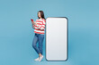 © ViDi Studio - Full body young woman she wear red striped sweatshirt casual clothes stand near big huge blank screen mobile cell phone with area using smartphone isolated on plain blue background. Lifestyle concept.