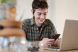 © Studio Marmellata - A happy young person with short hair smiles while using a smartphone. They are seated at a table with a laptop nearby, enjoying a casual moment of digital interaction.