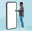 © Prostock-studio - Side View Of Black Guy Using Big Cellphone With Blank White Screen Touching Huge Display Panel With Finger, Cheerful Man Standing On Blue Background, Ordering Food Delivery, Mock Up. Full Body Length