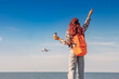 © EdNurg - Young woman passenger with backpack holding coffee cup, raising arm with peace sign, looking at flying aircraft